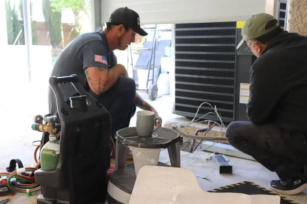 Two men work on a HVAC system in a garage, with tools and equipment around them. A cup sits on a small table nearby.